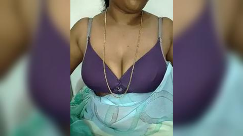 Media: Video of a woman with medium-dark skin, wearing a purple bra and a light blue floral-patterned garment. The image is slightly blurred, with focus on her chest and upper torso.