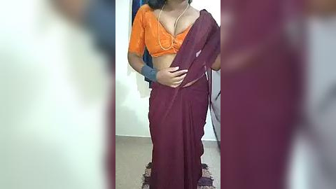 Media: Video of a woman in an orange blouse and maroon sari, standing in a room with beige carpet, blurry background, and indistinct figures.