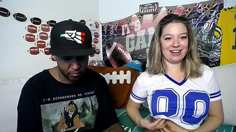 Media: Video of a young black man in a black hat and shirt, sitting next to a white woman with blonde hair in a white jersey. Background features sports memorabilia and a \"FAN\" banner.