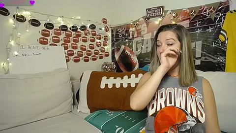 Media: Video of a young woman with shoulder-length brown hair, wearing a \"Browns\" T-shirt, sitting on a beige couch in a cozy living room decorated with football-themed posters and string lights.