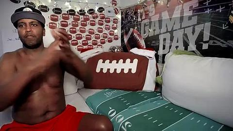 Media: Video of shirtless Black man in red shorts, sitting on a bed covered with green and white football-themed bedding, surrounded by football-themed decorations.