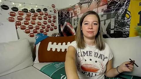 Media: Video of a young woman with straight brown hair, wearing a white t-shirt with a football design, seated on a beige sofa. Background features football-themed decor, including a large football pillow and framed football posters.