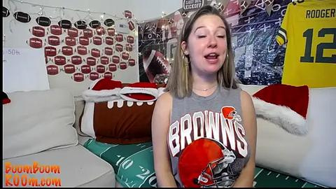 Media: Video of a young Caucasian woman with light skin, shoulder-length blonde hair, and a sleeveless \"Browns\" football jersey, sitting on a couch adorned with football-themed pillows and decor.