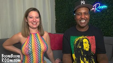 Media: Video of a white woman with straight brown hair in a colorful, high-cut, sleeveless dress, and a Black man with dreadlocks in a black t-shirt with a Bob Marley graphic, both smiling, in a room with greenery and a New York logo.