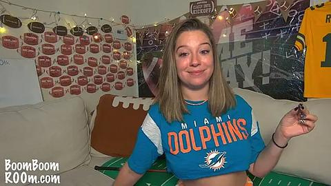 Media: A video of a young woman with straight, shoulder-length blonde hair, wearing a blue Miami Dolphins football jersey, posing in a living room decorated with football-themed decor, including a large \"GO DOLPHINS\" banner and string lights.