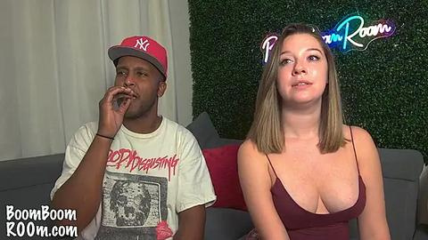 Media: Video of a young, light-skinned woman with straight, shoulder-length blonde hair wearing a low-cut, maroon tank top and a \"Pandora Room\" headband. She sits next to a dark-skinned man in a white T-shirt with a red hat, both indoors.