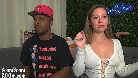 Media: Video of a Black man in a red cap and a black T-shirt with a political slogan, and a white woman in a white top, both seated on a couch in a room with a green wall and a neon sign.