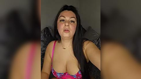 Media: Video of a curvy woman with long black hair, wearing a pink and blue floral bra, sitting on a dark couch, blurry background.