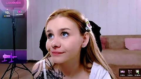 Media: Video of a young woman with fair skin, light brown hair adorned with a floral hairpin, wearing a white top, sitting in a living room with a beige couch and purple furniture, looking at a camera.