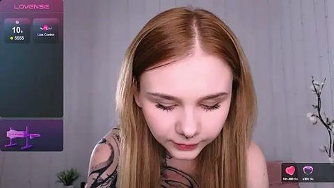 Media: Video of a young, fair-skinned woman with long, straight, light brown hair, wearing a black and white floral top, looking downward with closed eyes. Background features a grey wooden wall, a pink heart icon, and a pink \"Love\" text.