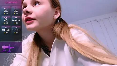 Media: Video of a fair-skinned, blonde-haired woman with pigtails, wearing a white lab coat, looking contemplative. Background features a white wall with a string of fairy lights and a purple digital interface.