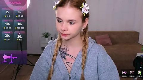 Media: Video of a young Caucasian woman with fair skin, light brown hair, and a floral headband, wearing a gray hoodie, in a modern living room with a beige sofa.