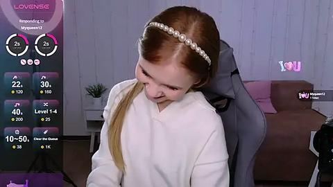 Media: A video of a young, fair-skinned girl with straight, long red hair, wearing a white headband and a white sweater, seated on a grey gaming chair in a modern living room.