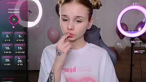 Media: Video of a young girl with light skin, blonde pigtails, wearing a white T-shirt, sitting in a room with a ring light, balloons, and a computer screen displaying \"Lovely\" and \"Recovery\" apps.