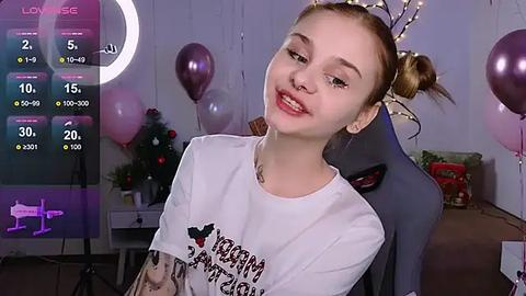 Media: Video of a young, fair-skinned woman with blonde pigtails, smiling in a cozy room with Christmas decorations, wearing a white T-shirt with a graphic.