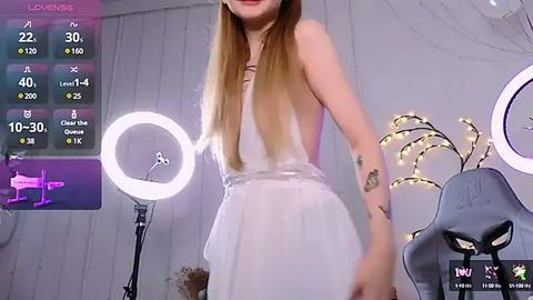 Media: A video of a slender, light-skinned woman with long blonde hair, wearing a white sleeveless dress, standing in front of a glowing ring light and a gray gaming chair.