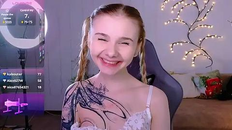 Media: Video of a smiling young woman with fair skin, brown pigtails, wearing a white lace bralette, showing a large black tattoo on her right shoulder. Background includes a lit Christmas tree, a gaming chair, and a soft-focus room with a pink ring light.