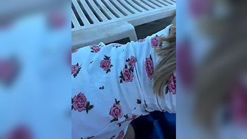 Media: Video of a woman with blonde hair, wearing a white blouse with pink rose pattern, lying on a white grate, her head to the left. Background shows a blurry, white building.