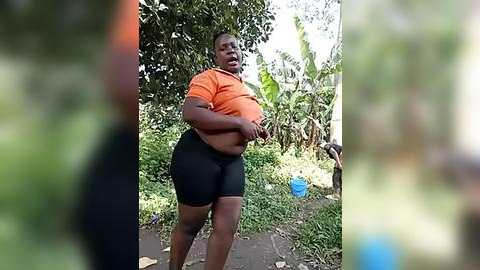 Media: Video of a plus-size Black woman with dark skin, wearing an orange top and black shorts, standing outdoors amidst lush greenery, holding her large breasts.