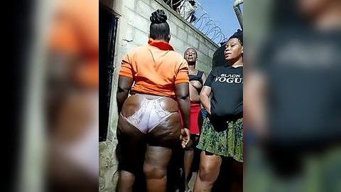 Media: Video of four Black women walking outdoors, one in an orange shirt and white lace thong, another in a green skirt, another in a black shirt, and a fourth in a red top. They pass a concrete wall with barbed wire.