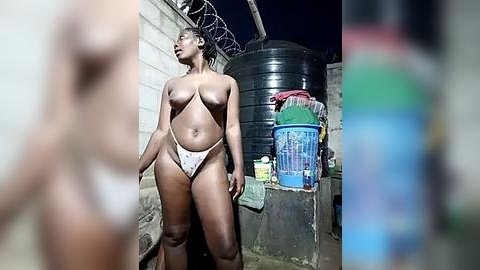 Media: Video of a topless, dark-skinned woman with medium-sized breasts, wearing only a white thong, standing in a dimly-lit, urban alley. Concrete walls, black water tanks, and colorful plastic buckets are in the background.