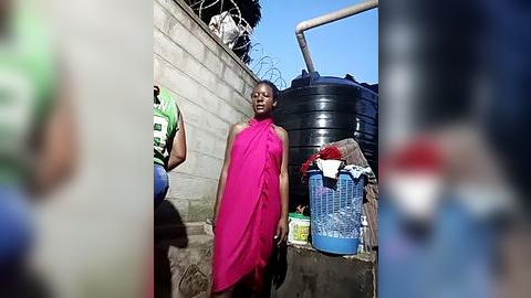 Media: Video of a woman in a bright pink dress standing near a large black water tank and laundry basket in a gritty urban setting with a wire fence and concrete wall.