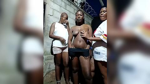 Media: Video of three African women standing in front of a concrete wall at night, wearing white tank tops and shorts. One woman is topless, exposing her breasts, with a hand covering her abdomen.