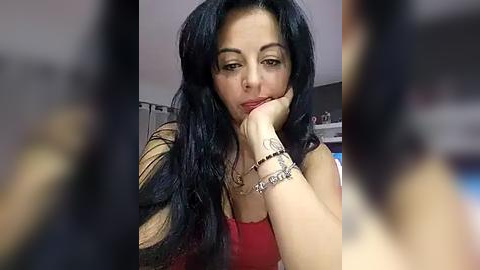 Media: Video of a woman with long black hair, fair skin, and a red top, sitting pensively with her chin resting on her hand, indoors with a blurred background.