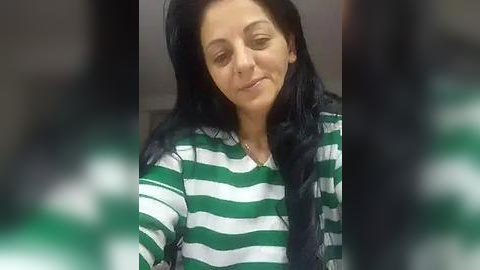 Media: Video of a young woman with long black hair, wearing a green and white striped shirt, smiling slightly. Background is blurred, possibly indoors.