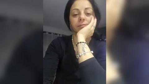 Media: Video of a woman with short black hair, resting her chin on her hand, wearing a black sweater, silver bracelet, and a tattoo on her wrist. Blurry background.