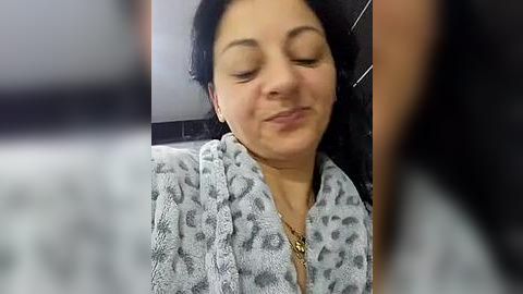 Media: Video of a middle-aged woman with medium skin tone, wearing a leopard-print robe, smiling with closed eyes in a bathroom with black and white tiled walls.