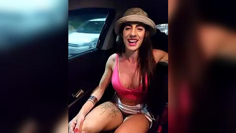 Media: Video of a smiling, tanned woman with long dark hair, wearing a pink tank top, denim shorts, and a tan hat, sitting in a car at night.