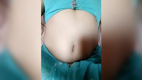 Media: Video of a pregnant woman's belly, wearing a teal blouse, with a visible navel and slight belly bulge. Background is blurred, focusing on the midsection.