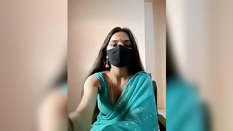 Media: Video of a South Asian woman with long black hair, wearing a turquoise sari and black face mask, seated indoors with blurred background.