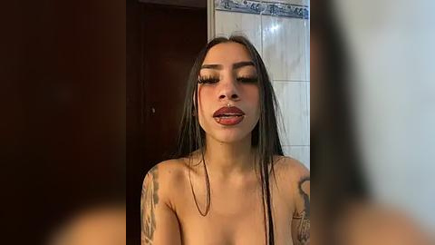 Media: A video of a young Latina woman with long, straight dark hair, wearing minimal makeup, and displaying tattoos on her arms, standing topless in a bathroom with tiled walls and a wooden door.