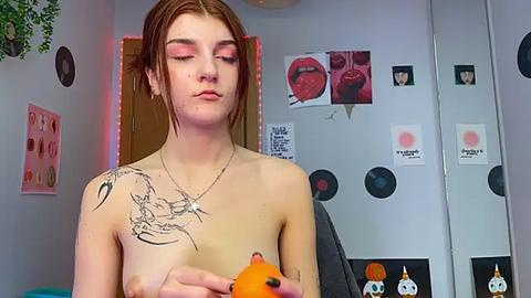 Media: Video of a young woman with pale skin, red hair, and pink eyeshadow, topless, holding a plush fox. Background features posters and records, creating a casual, artistic ambiance.