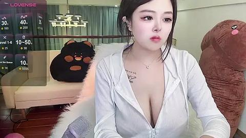 Media: A video of an Asian woman with fair skin, dark hair, and a low-cut white zip-up top, seated in a modern living room with plush brown furniture, a bear plush, and a digital display.