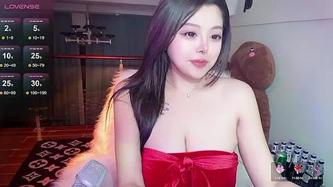 Media: Video of a young Asian woman with long black hair, wearing a strapless red dress, seated in a cozy room with a plush teddy bear, wooden floor, and gaming setup.