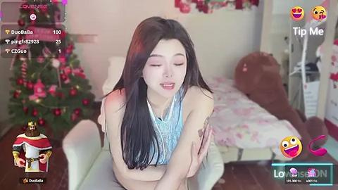 Media: Video of an Asian woman with long black hair, wearing a sleeveless blue top, sitting on a bed in a festive, decorated room with a Christmas tree.