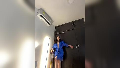 Media: Video of a woman in a blue robe, standing in a modern room with dark wooden cabinets, an air conditioner, and bright lighting.