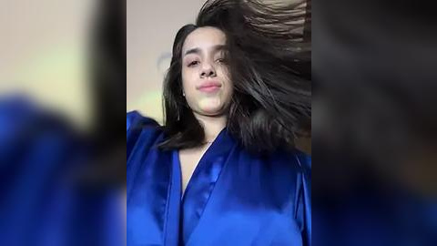 Media: Video of a woman with long, dark hair, wearing a blue robe, her hair flying, in a blurred, indoor setting with muted colors.