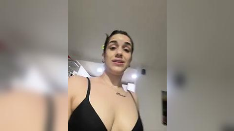 Media: A video of a young woman with light skin, dark hair tied back, wearing a black bra, smiling, with a small tattoo on her left shoulder, standing in a dimly lit room with beige walls.