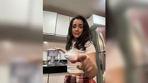 Media: A video of a young woman with long black hair, wearing a white crop top and plaid skirt, standing in a modern kitchen, holding a spoon towards the camera.