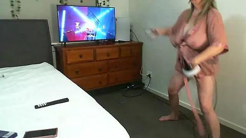 Media: Video of a woman in a pink robe, barefoot, energetically playing a video game on a TV stand in a bedroom with a bed, dresser, and a wall-mounted TV.