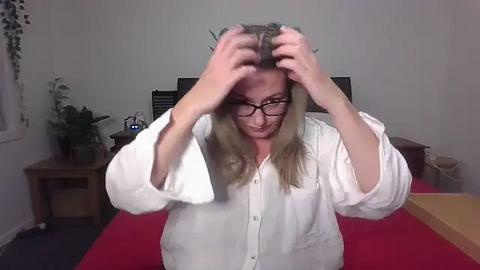 Media: Video of a blonde woman with glasses, wearing a white shirt, frantically combing her hair, seated at a desk with a red tablecloth, surrounded by potted plants and a monitor.