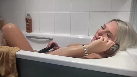 Media: A video of a nude blonde woman with a slender build, lying in a white bathtub with a black handheld showerhead, her right arm resting on the edge, water glistening on her skin, beige towel nearby, tiled white bathroom background.