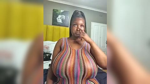 Media: A video of a plus-sized Black woman with dark skin and large breasts, wearing a colorful, striped tank top, sitting on a bed with a yellow headboard, in a dimly lit room.