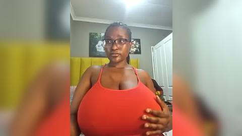 Media: A video of a dark-skinned woman with a large bust, wearing a tight red tank top, standing in a bedroom with a yellow upholstered headboard and framed artwork.