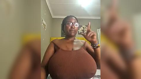 Media: Video of a dark-skinned woman with large breasts, wearing glasses, a brown ribbed top, and bracelets, making a peace sign, in a dimly lit room with beige walls.