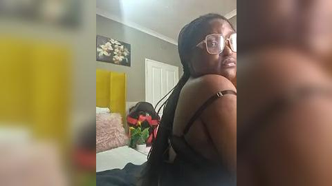 Media: Video of a young Black woman with medium skin tone, wearing glasses and black bra, standing in a bedroom with yellow curtains, white door, and floral painting on the wall.